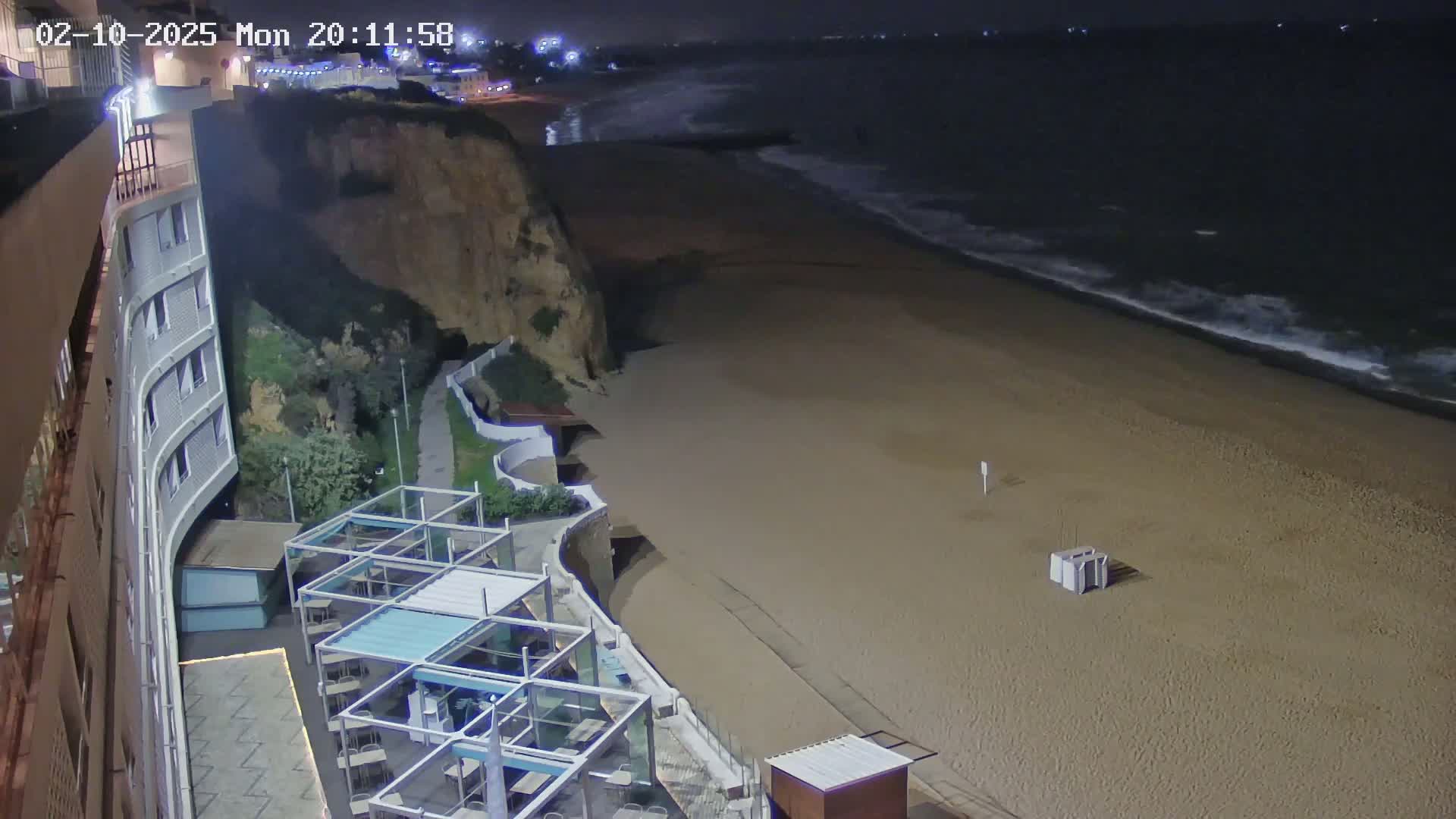 A nighttime beach scene shows a sandy shore with gentle waves, a cliff with buildings and outdoor seating areas, all under a dark sky.