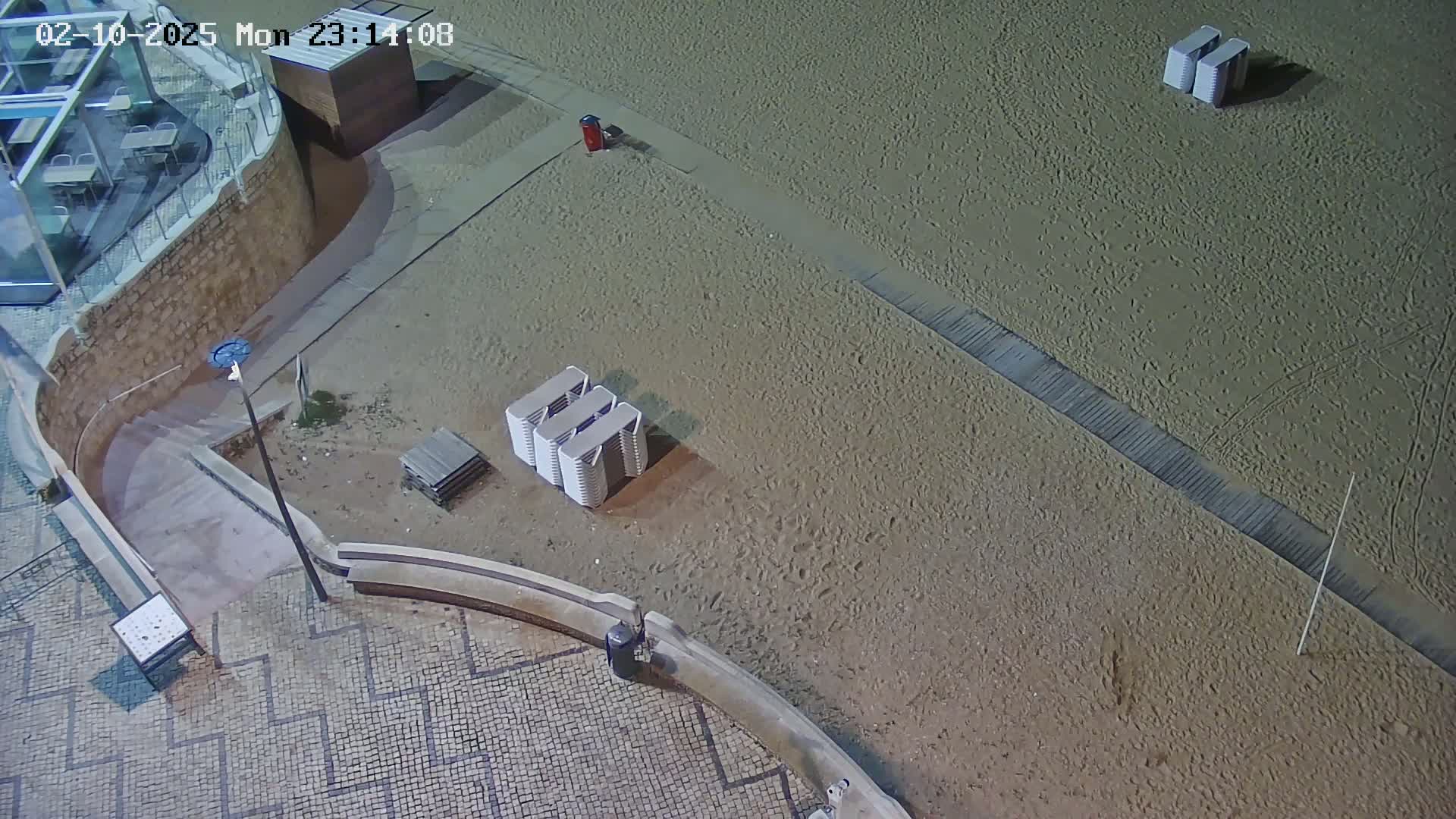 A mostly empty sandy beach at night is viewed from above, showing stacked beach chairs and a walkway leading to a building.