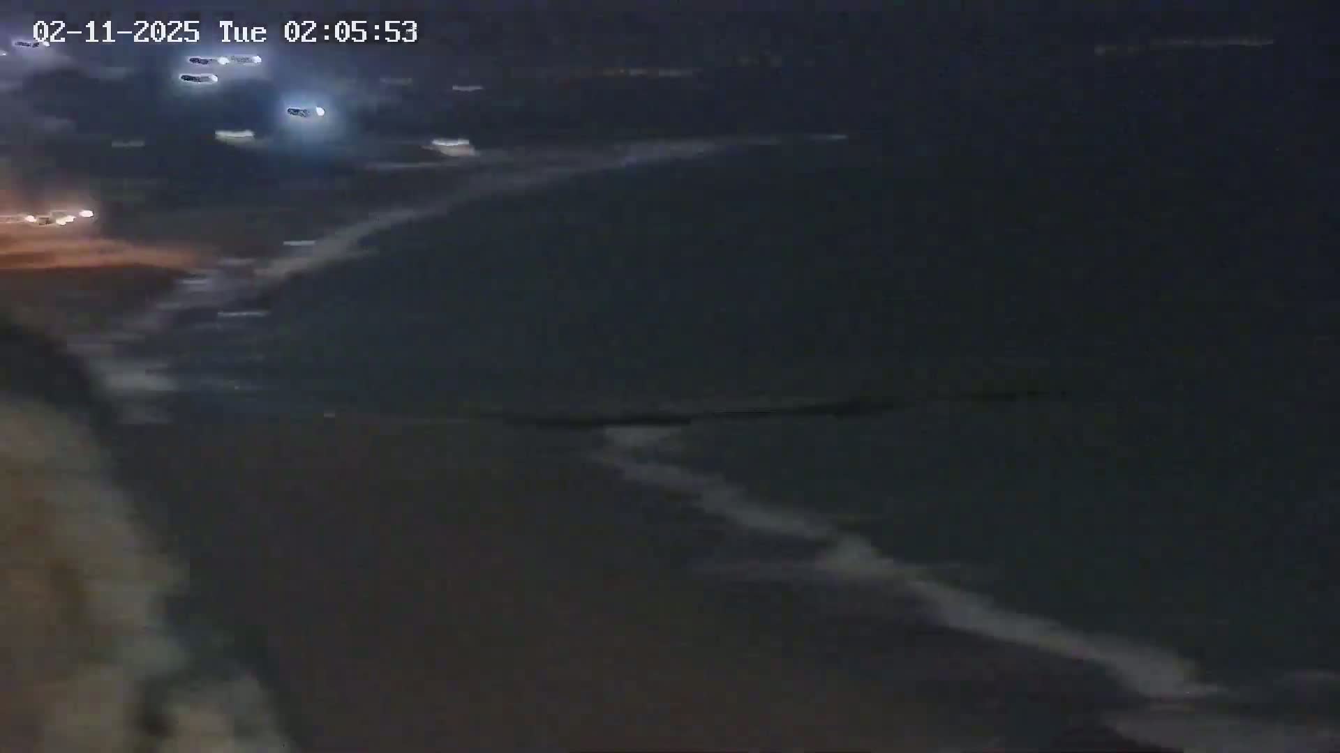 A dark, nighttime beach scene shows gentle waves lapping at the shore, with a few distant car lights visible along a road.