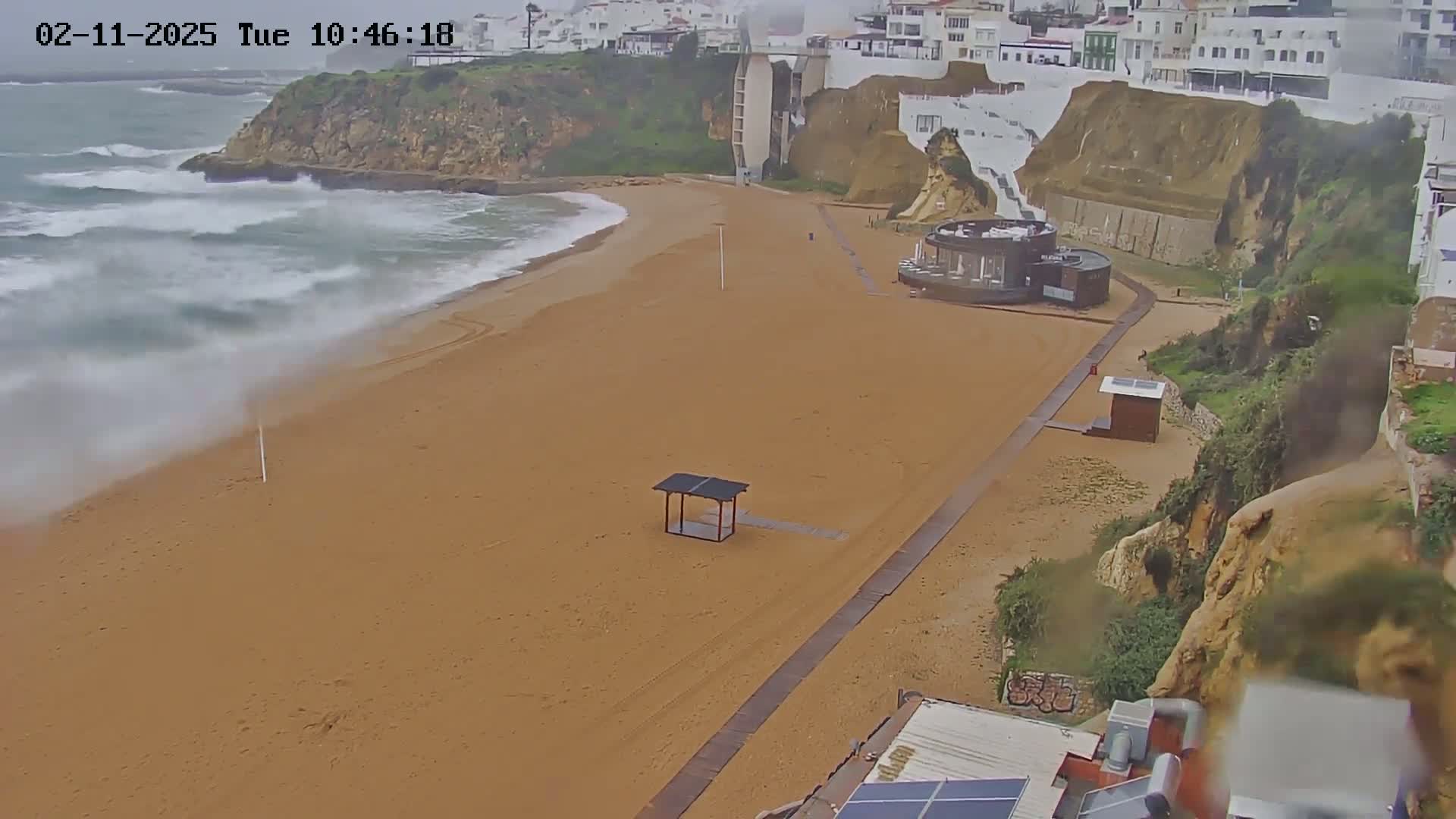 A sandy beach with a boardwalk and small structures is seen from above, with choppy waves crashing on the shore under overcast skies.