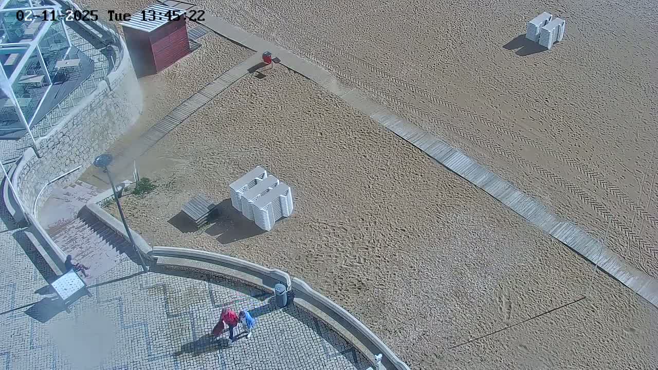 A sandy beach with boardwalk paths, stacked beach chairs, and a few people near a stone staircase leading to a building.