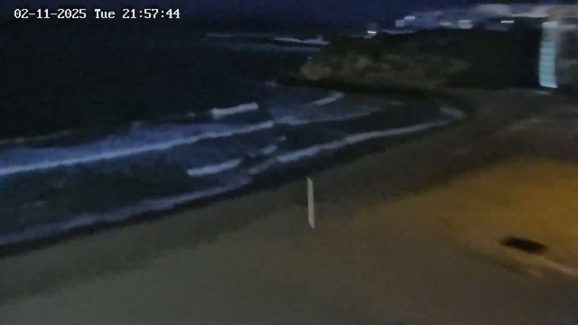 Dark, nighttime waves crash on a shore near a road.
