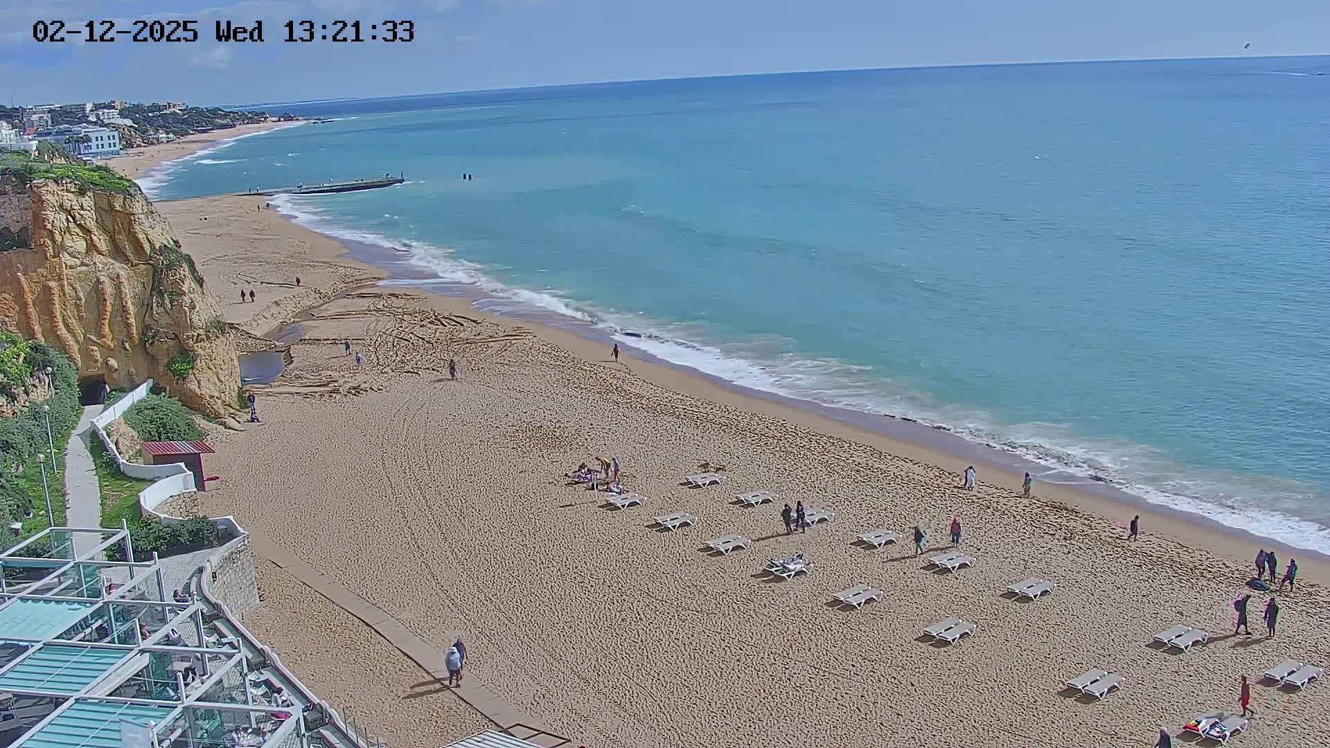 A sandy beach with a few people and lounge chairs stretches along a clear, blue ocean under a sunny sky.