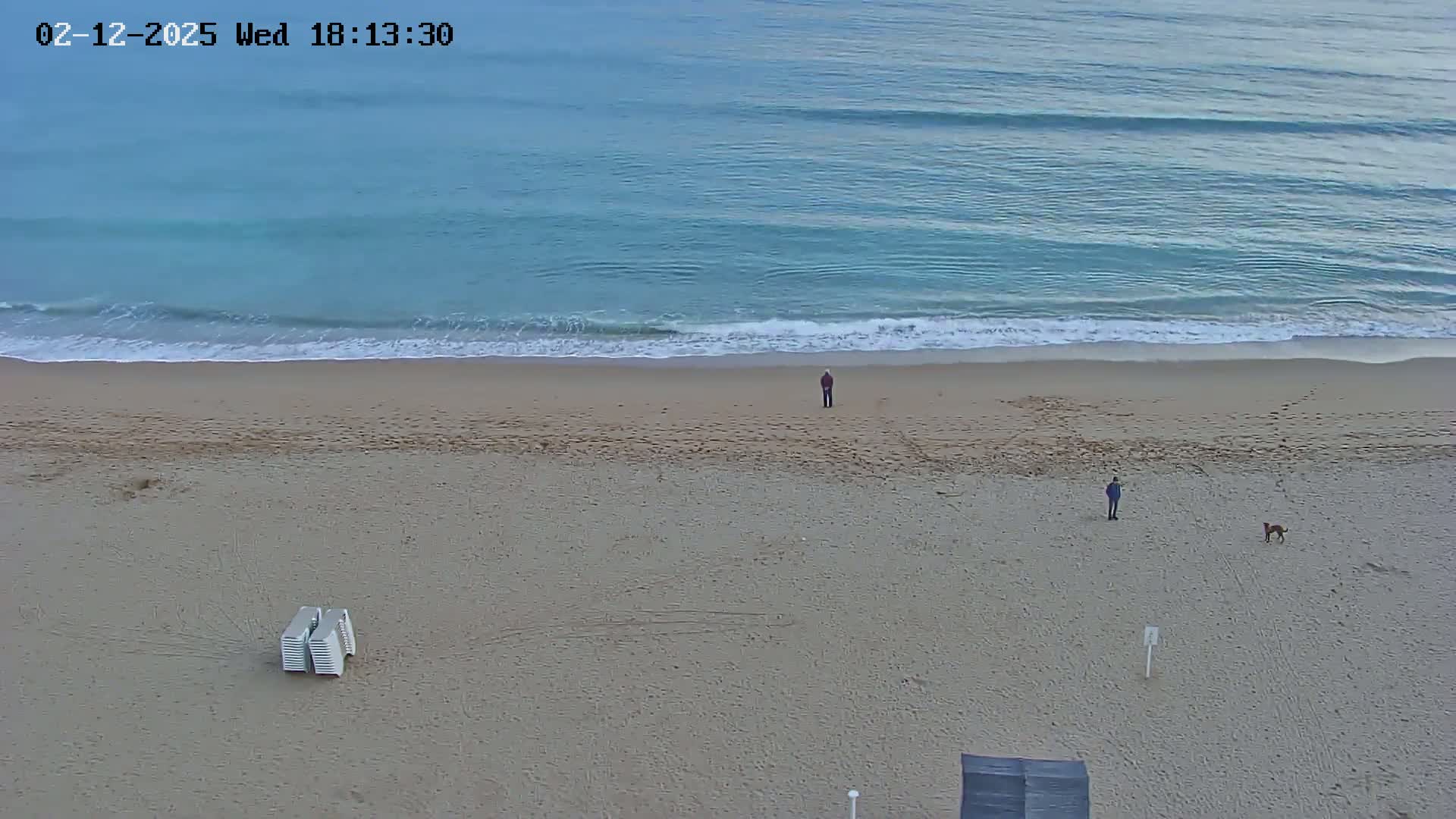 A sandy beach with gentle waves and calm water, a few people and a dog, and a stack of beach chairs are visible.