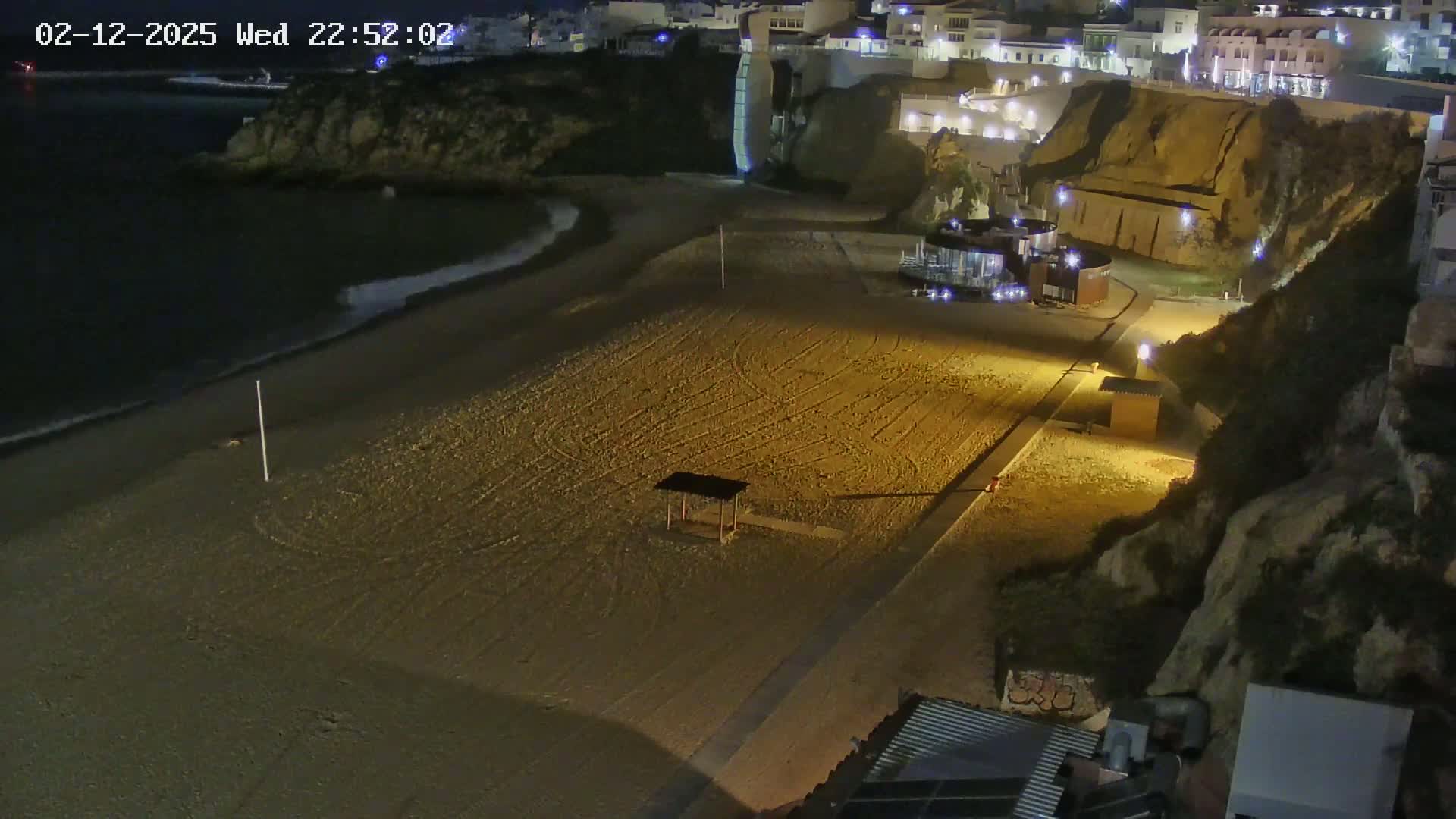A mostly empty sandy beach at night is illuminated by artificial lights, with calm waves lapping the shore and buildings perched on a cliff overlooking the scene.