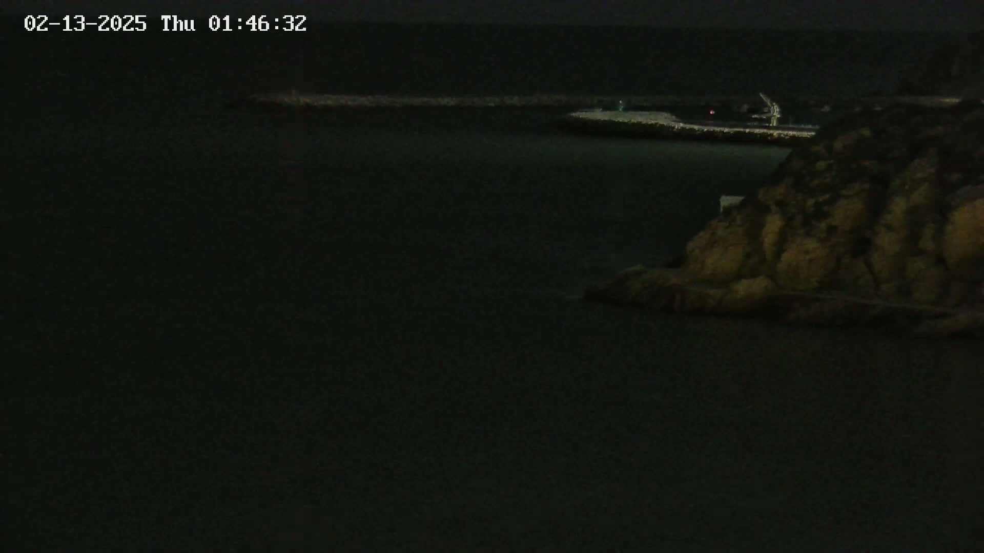 A dark, nighttime view of a body of water with a rocky shoreline and a distant, faintly lit structure.