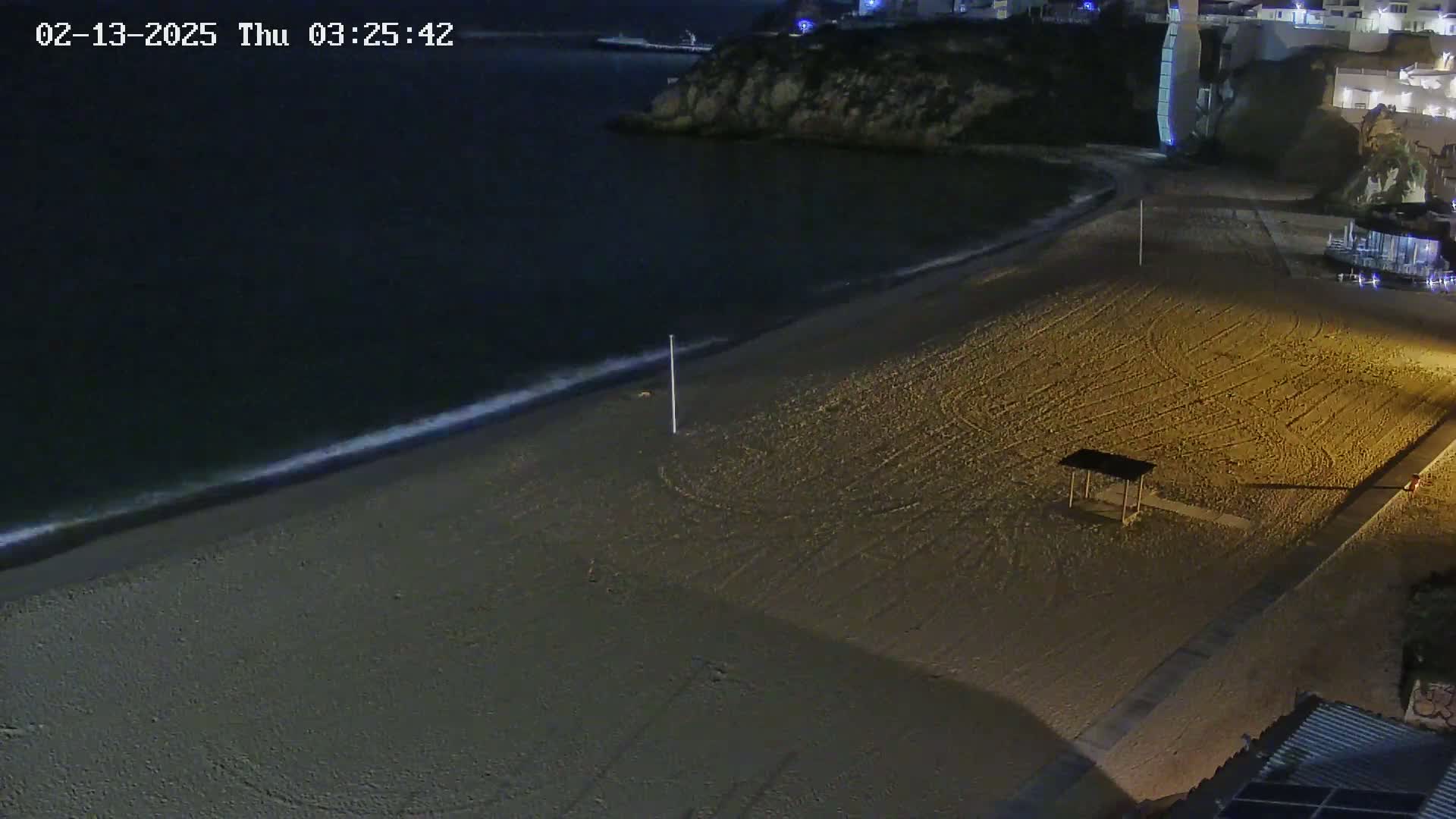 A sandy beach at night, illuminated, with gentle waves lapping the shore, and a small structure on the sand.