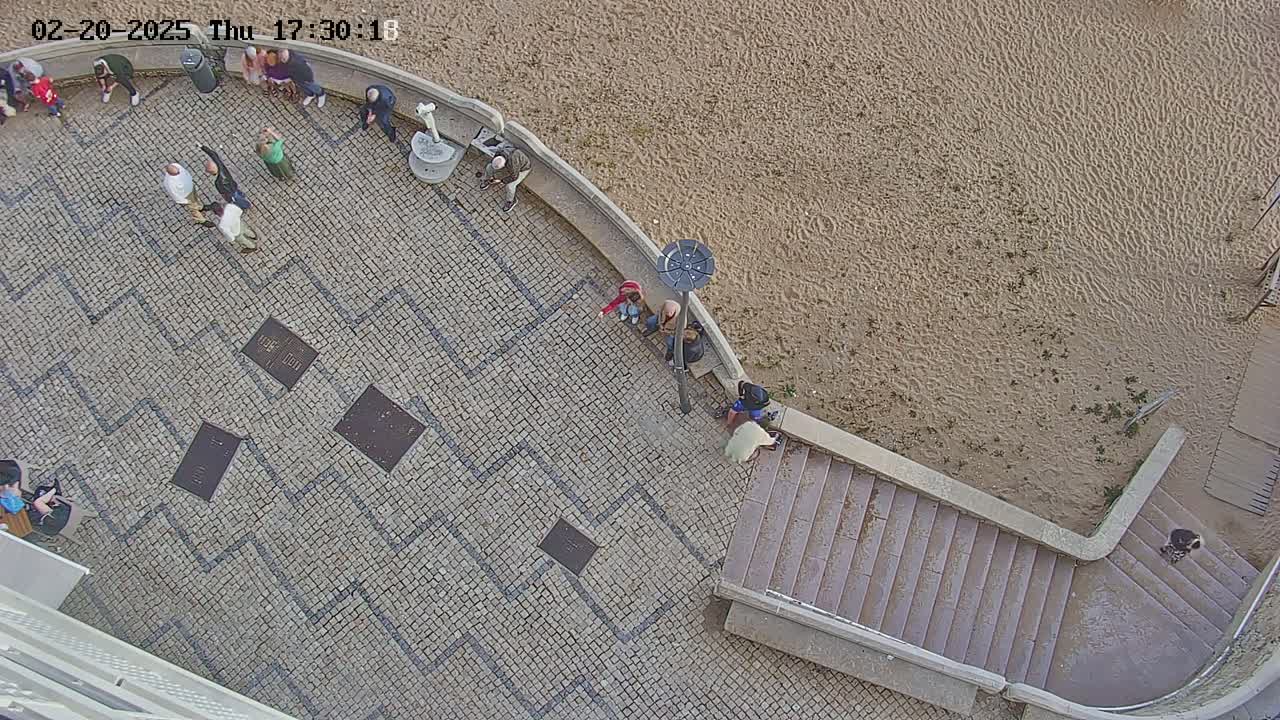 An aerial view shows a stone plaza with several people scattered around, leading down to a sandy beach.