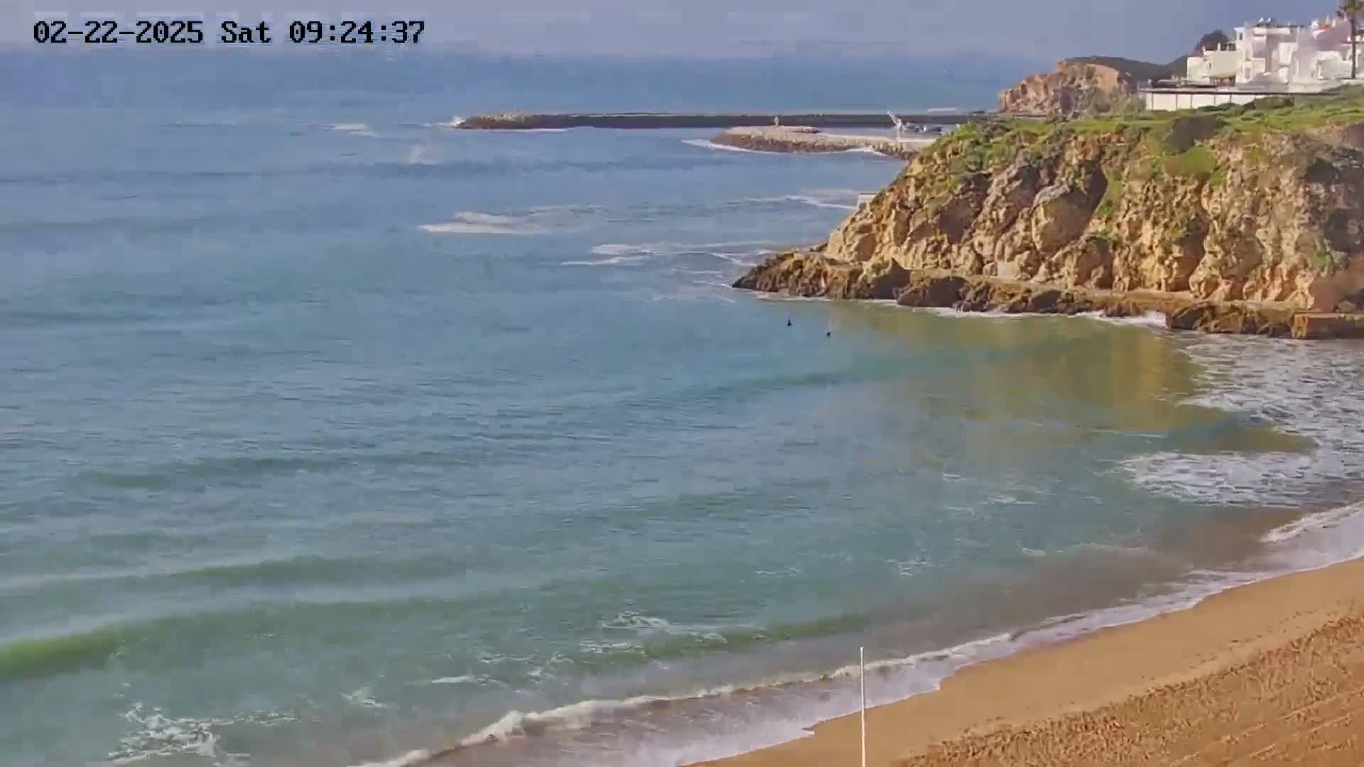 A sandy beach with calm, light-green ocean waves laps the shore near a rocky cliff on a sunny day.
