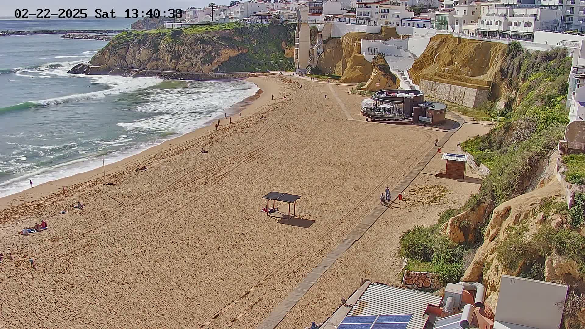 A sandy beach with a few people on it, next to a cliff with buildings on top, under sunny conditions with moderate waves in the ocean.