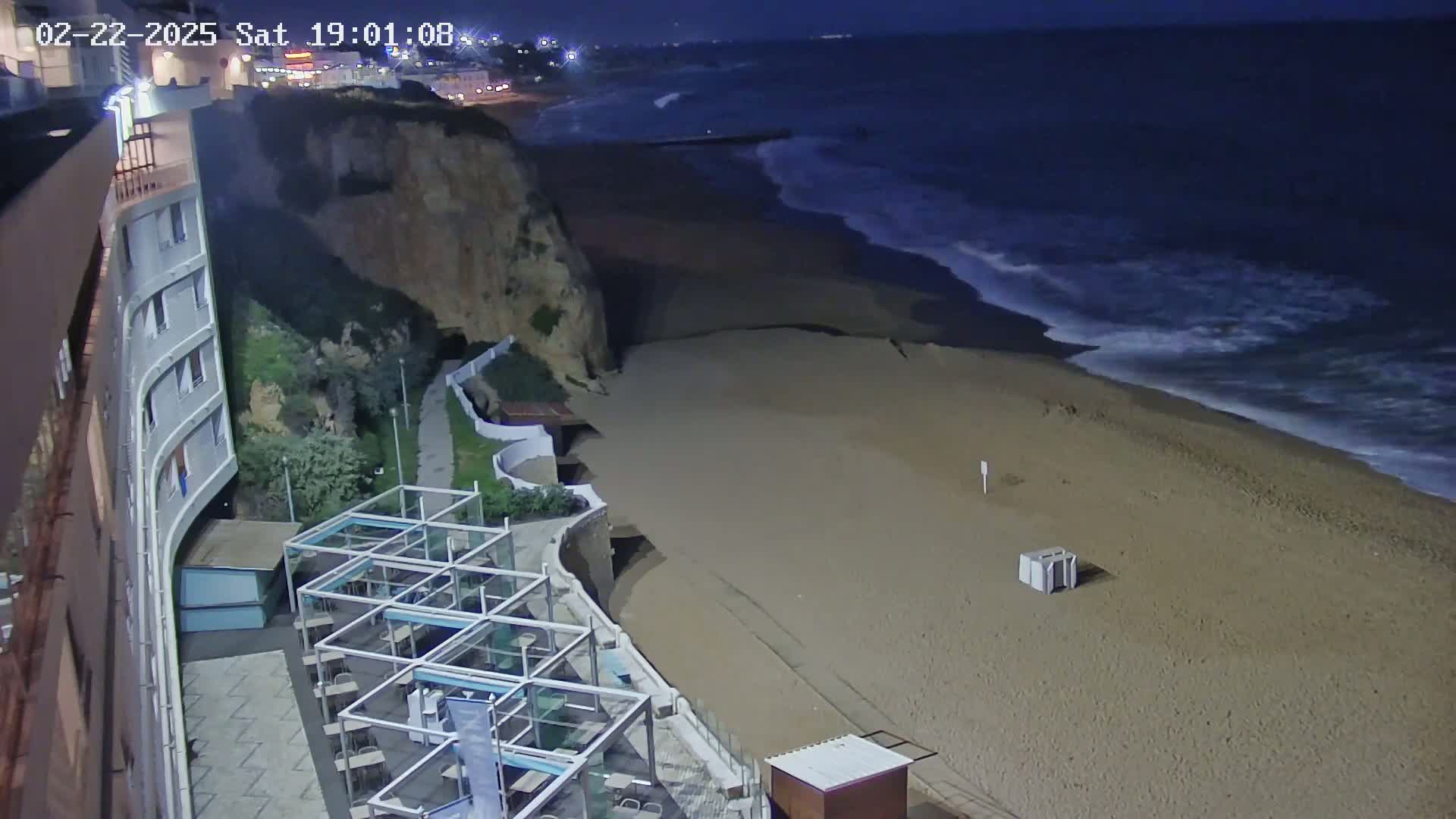 A nighttime view of a sandy beach with gentle waves under a dark sky, next to a cliffside building with outdoor seating.