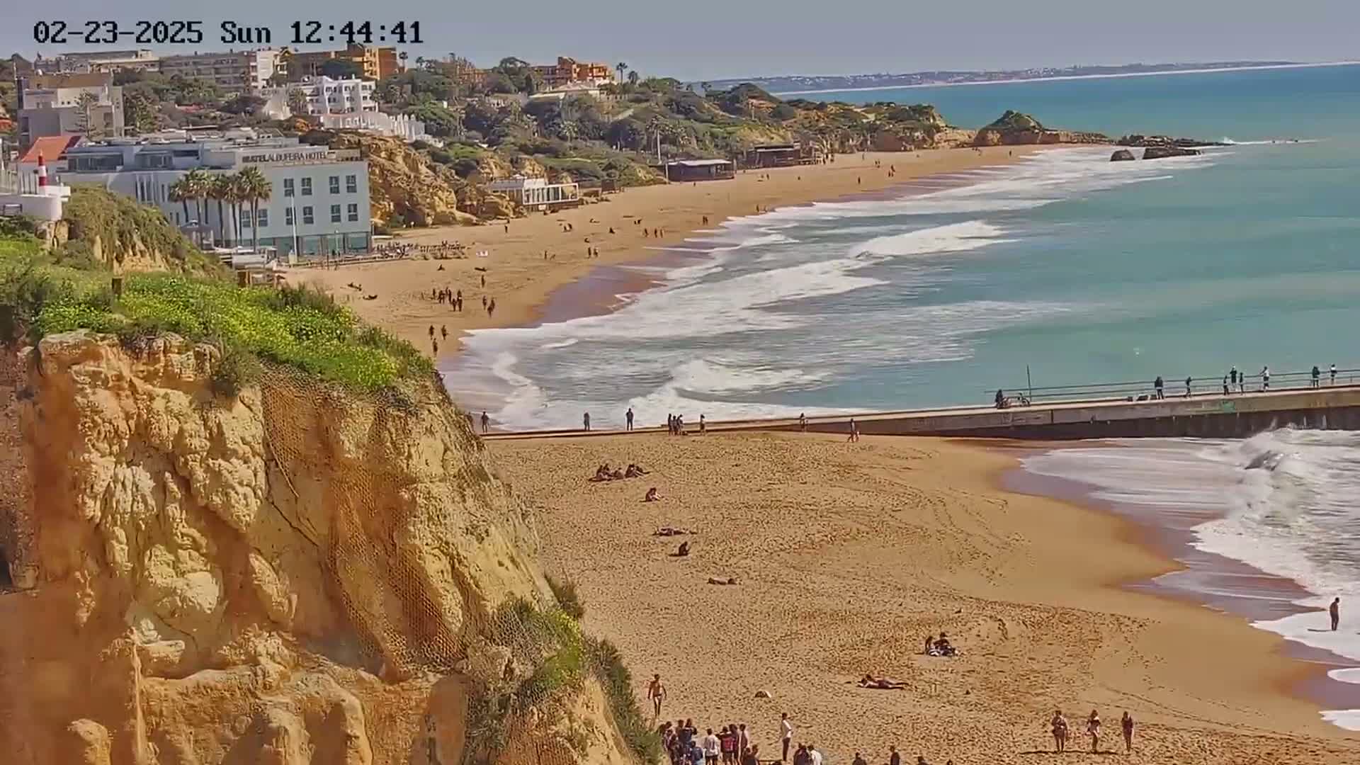 A sandy beach with moderate waves under a sunny sky is viewed from a cliff overlooking a boardwalk and buildings.