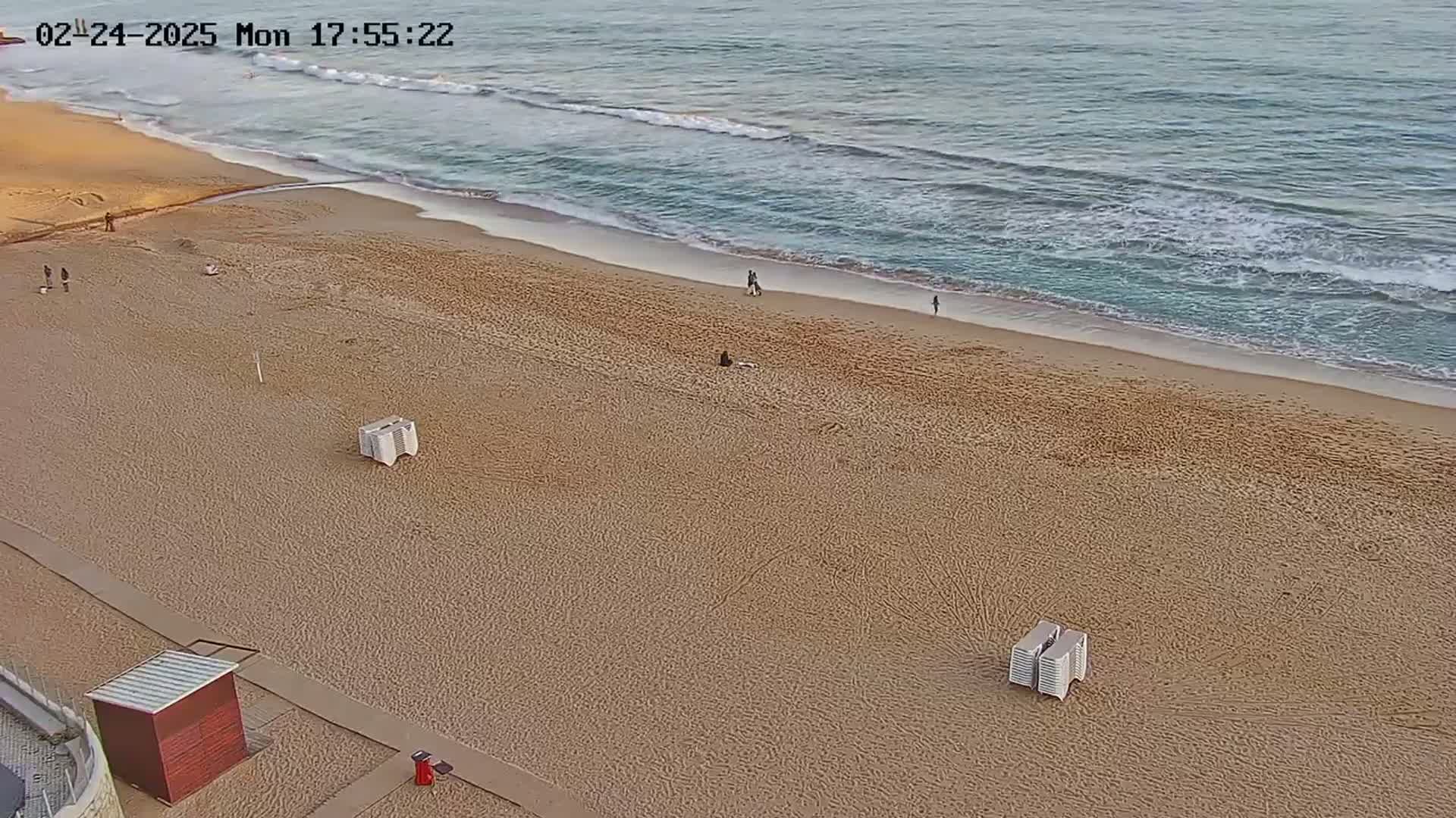 An expansive sandy beach with a few people scattered along it and gentle waves lapping the shore on a seemingly calm day.