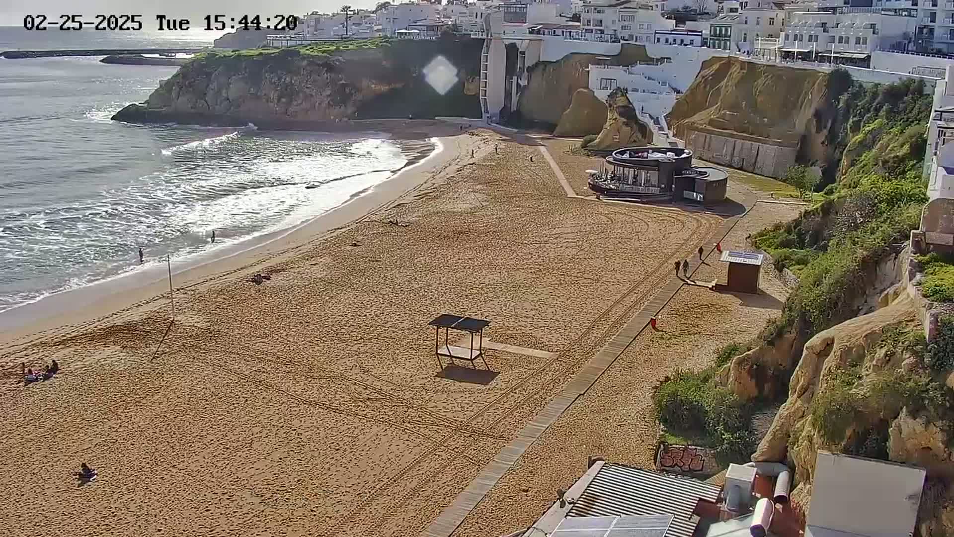A sandy beach with a few people is situated below a cliffside town on a sunny day with gentle waves lapping the shore.