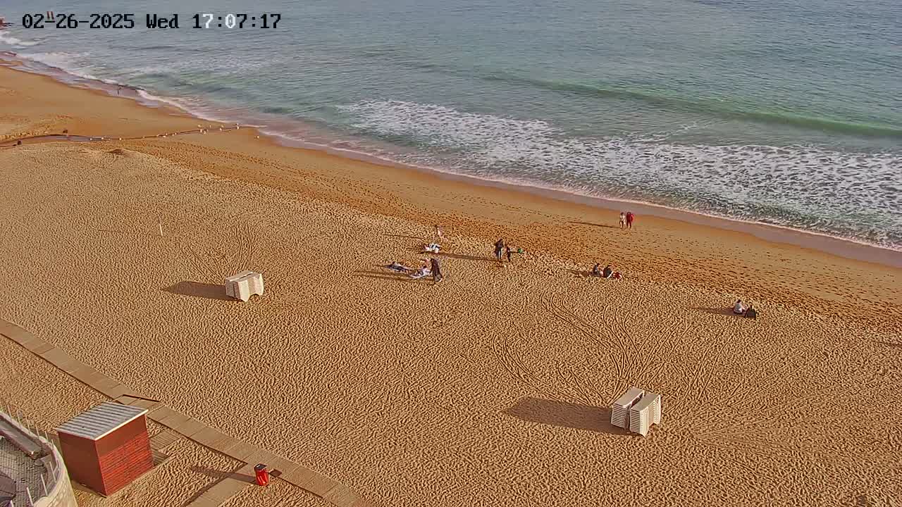 A sandy beach with a few people scattered across it, gently sloping to a calm ocean with small waves under a clear sky.