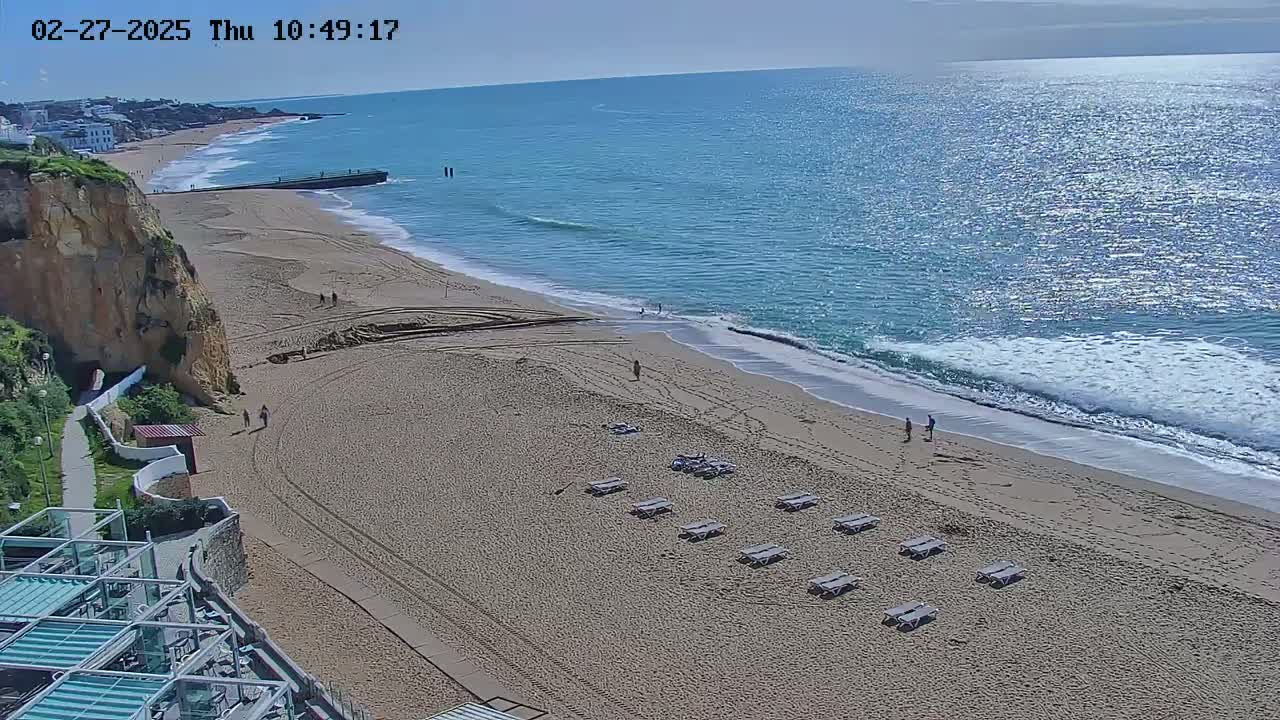 A sandy beach with a few people and lounge chairs stretches alongside a calm, blue ocean under a clear sky.