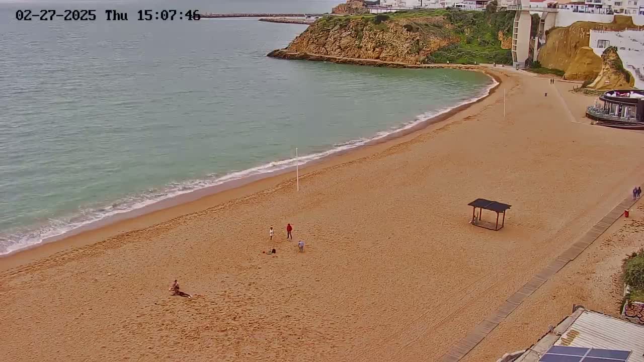 A sandy beach with a few people on it curves around a calm, light-green sea next to a cliffside with buildings.