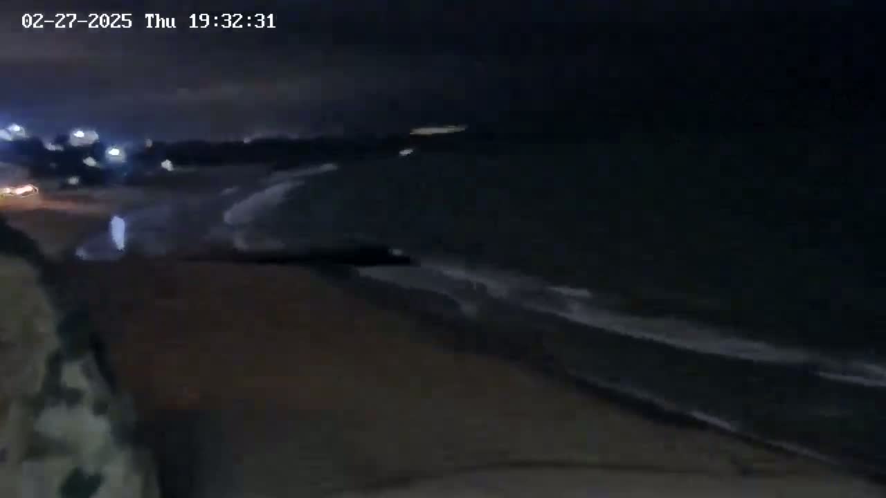A dark, nighttime beach scene shows gentle waves lapping the shore under a cloudy sky.