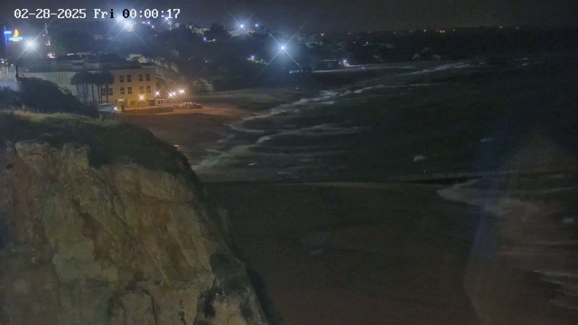 A nighttime view from a cliff overlooking a dark beach with gently lapping waves under a clear sky.