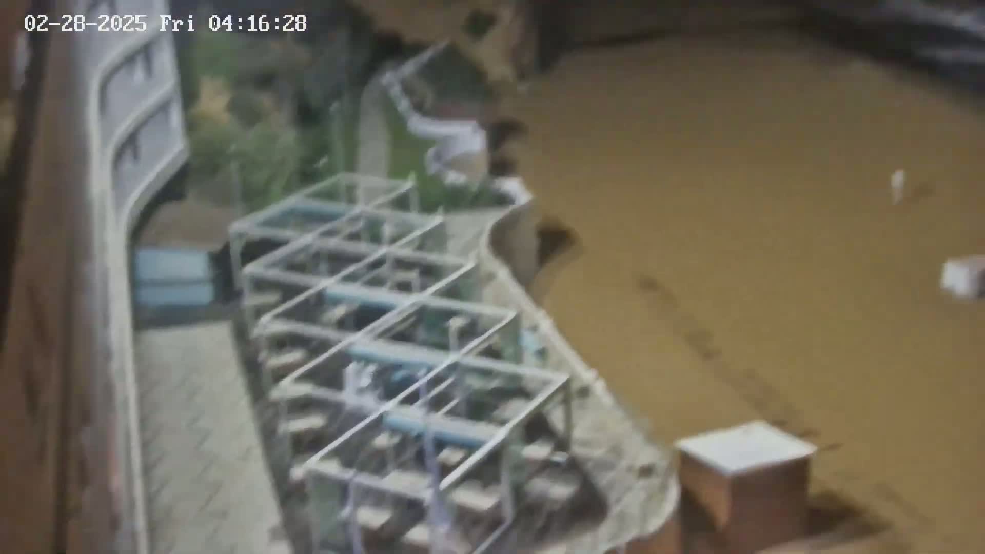 A high-angle, nighttime view shows muddy floodwaters surrounding a structure with several rows of caged equipment, possibly machinery.