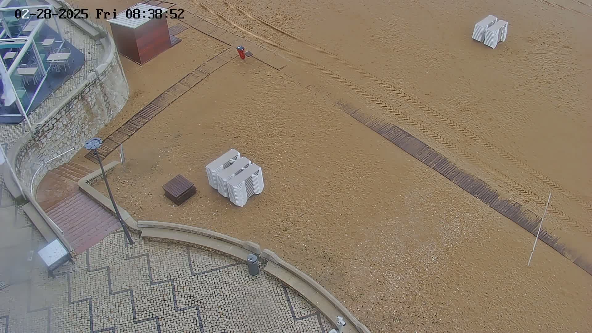 A mostly empty sandy beach is seen from above, with stacked beach chairs and tire tracks visible; a paved area with stairs leads to the beach from a building.