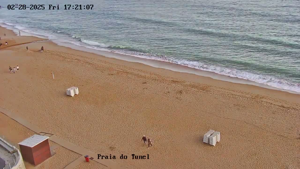 An overhead view shows a sandy beach with a calm ocean and a few people and beach chairs sparsely scattered across the sand.