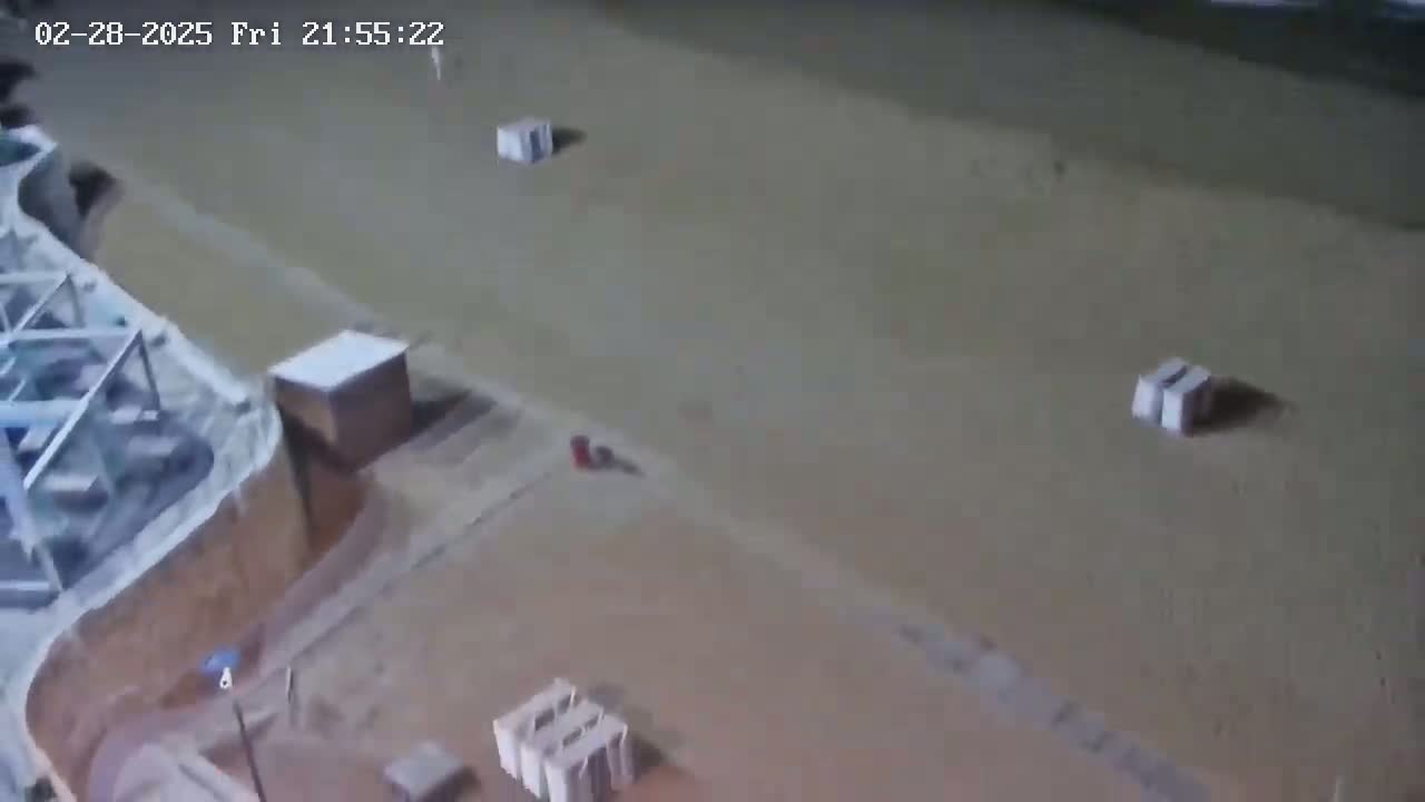 An aerial nighttime view shows a mostly empty sandy area with several small structures and a single small vehicle.