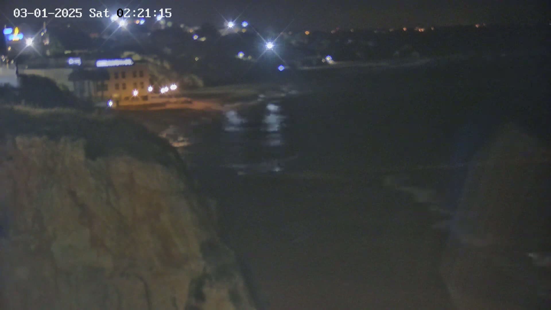 A nighttime view from a cliff overlooking a dark body of water, with a few lights visible on the opposite shore.