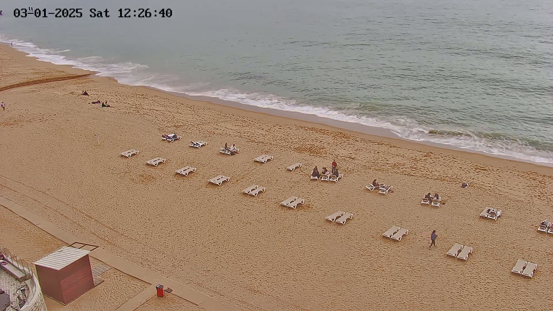 An overhead view shows a sandy beach with several people and lounge chairs, next to a calm, pale-green ocean under an overcast sky.