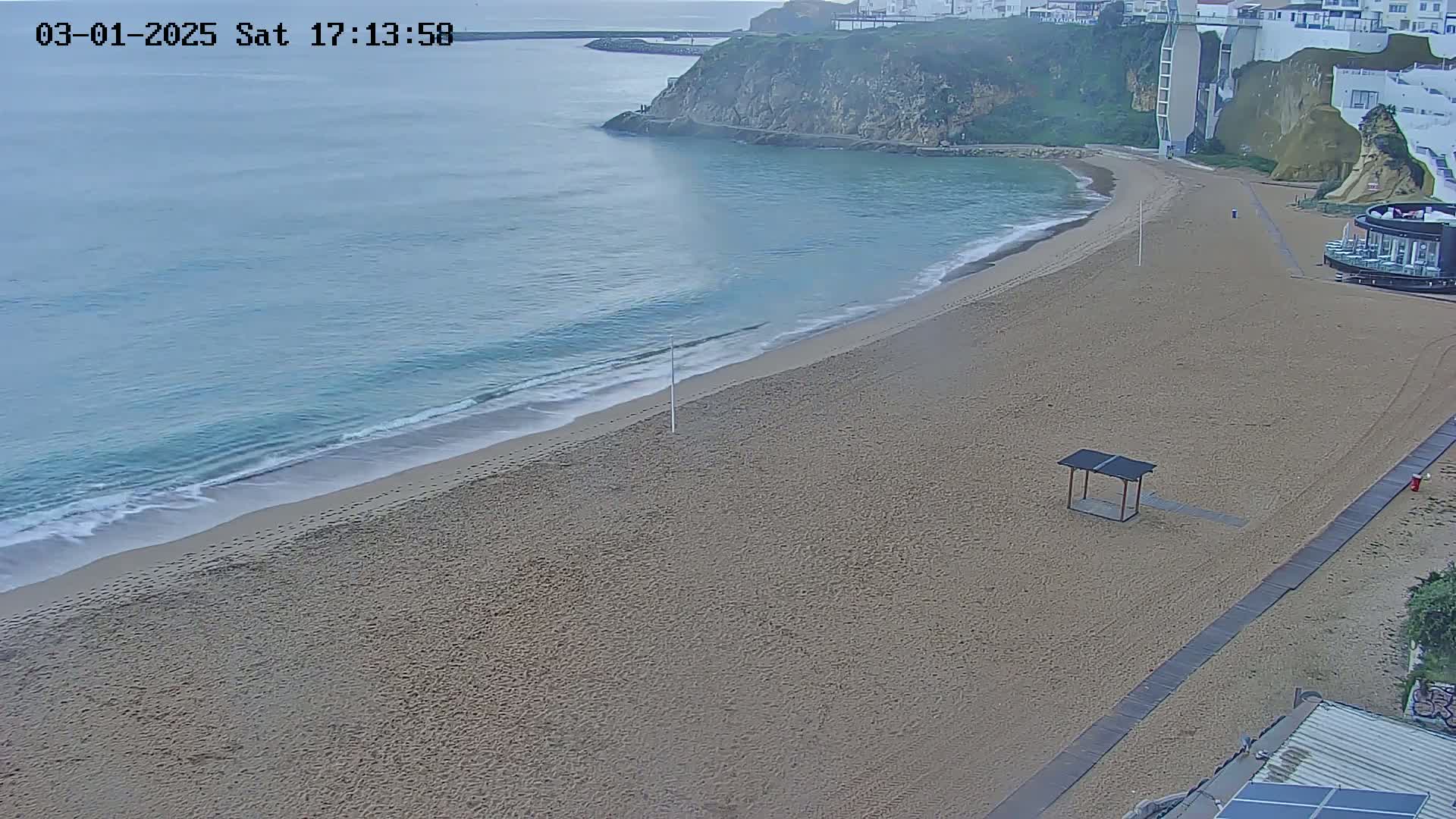 A mostly empty sandy beach curves alongside a calm, light-blue ocean under an overcast sky.