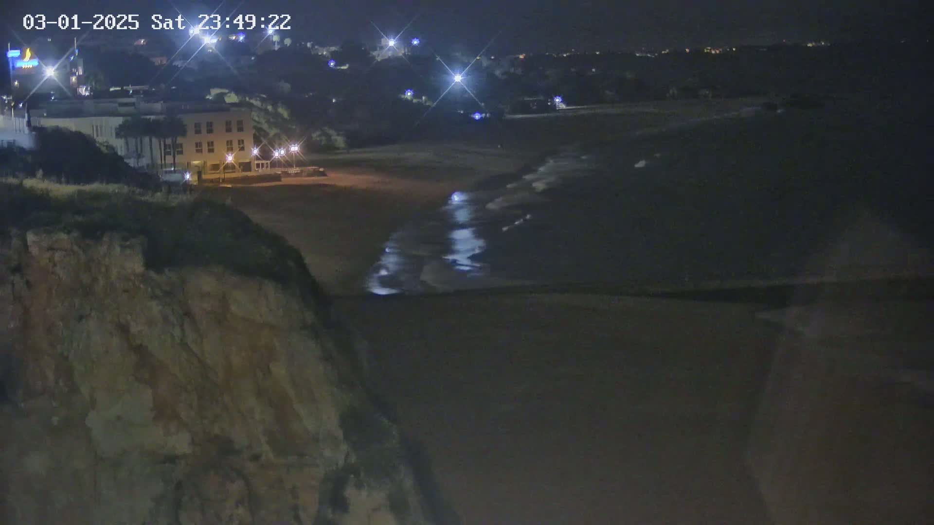 A nighttime view of a dark sandy beach with gentle waves lapping the shore, seen from a cliff overlooking a building and town lights.