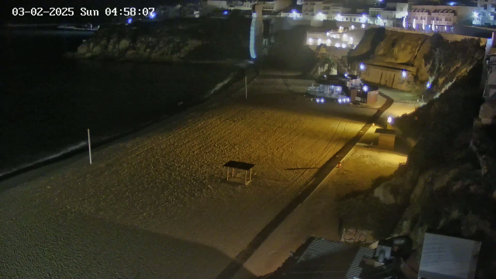 A mostly empty sandy beach at night is lit by artificial lights, with a calm dark sea in the background and buildings on a cliff overlooking the beach.