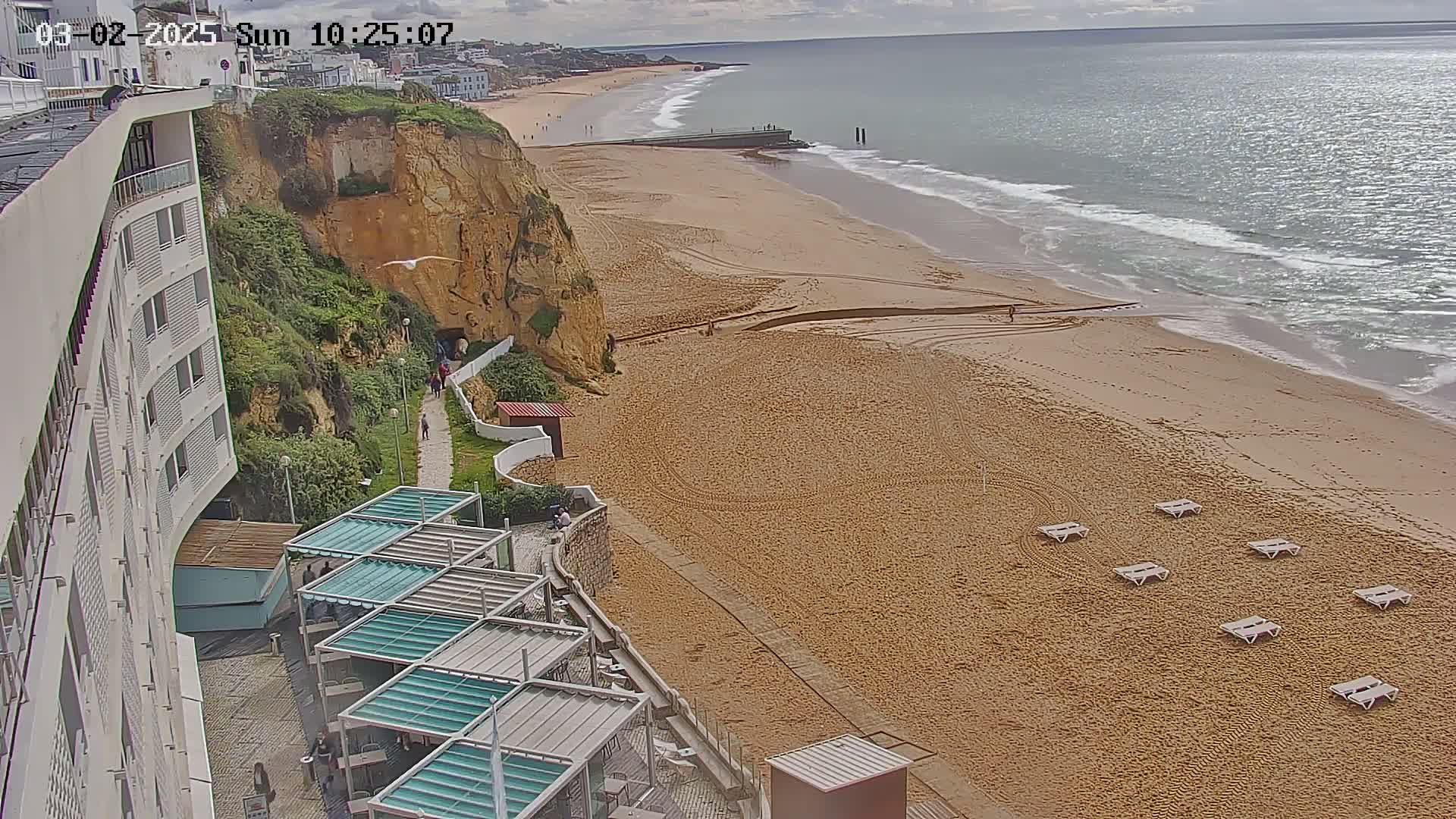 A sandy beach with several empty lounge chairs stretches out to a calm ocean under a partly cloudy sky, next to a cliff with a building on top.