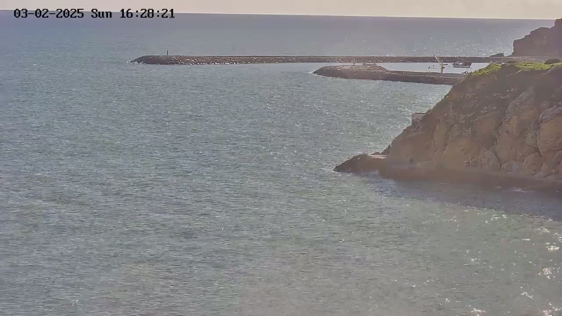 A rocky cliff overlooks a calm sea with a breakwater and a small harbor visible in the distance under a sunny sky.