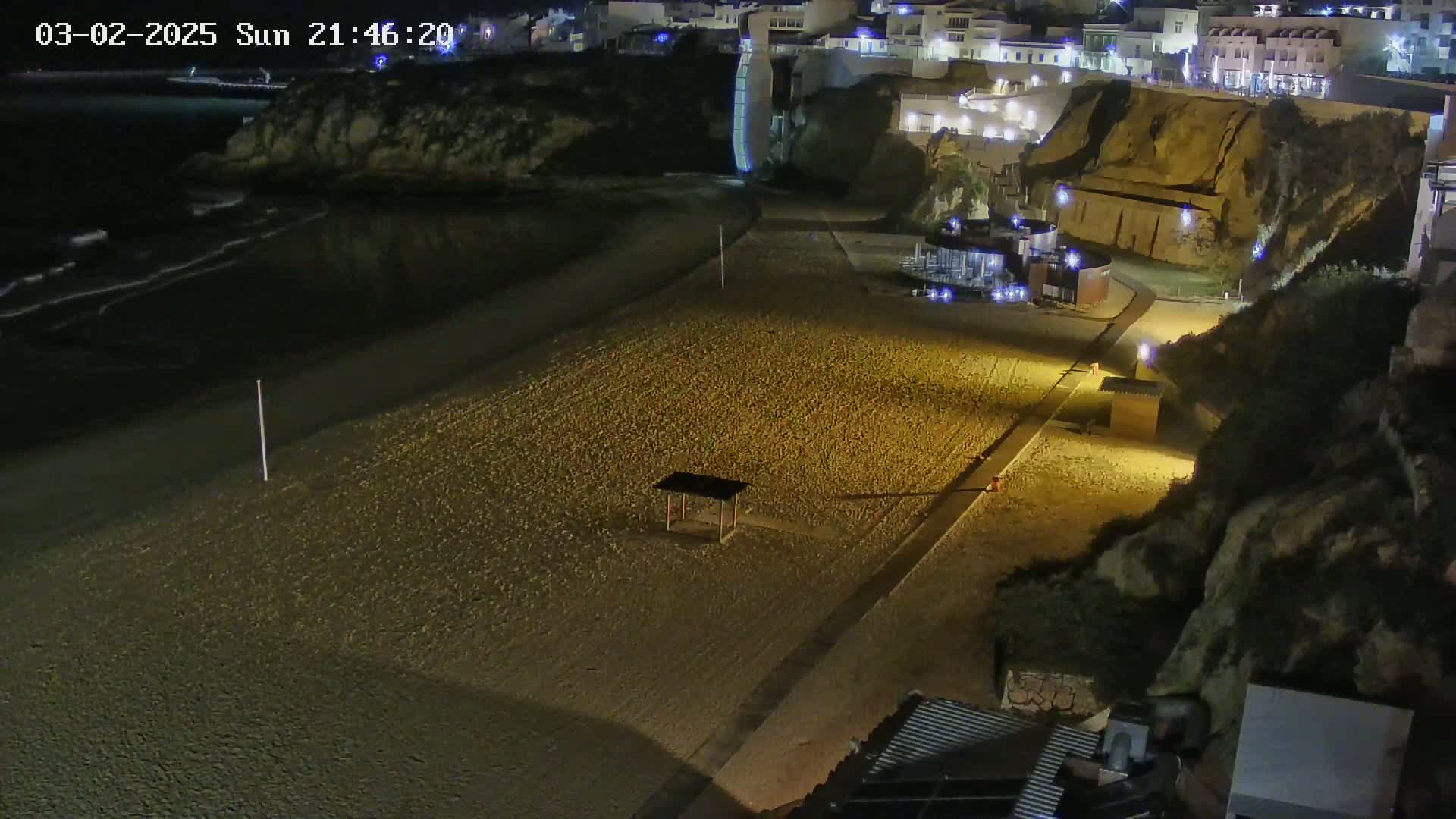 A sandy beach at night, illuminated by artificial lights, is bordered by a dark ocean with small waves and a cliff with buildings on top.