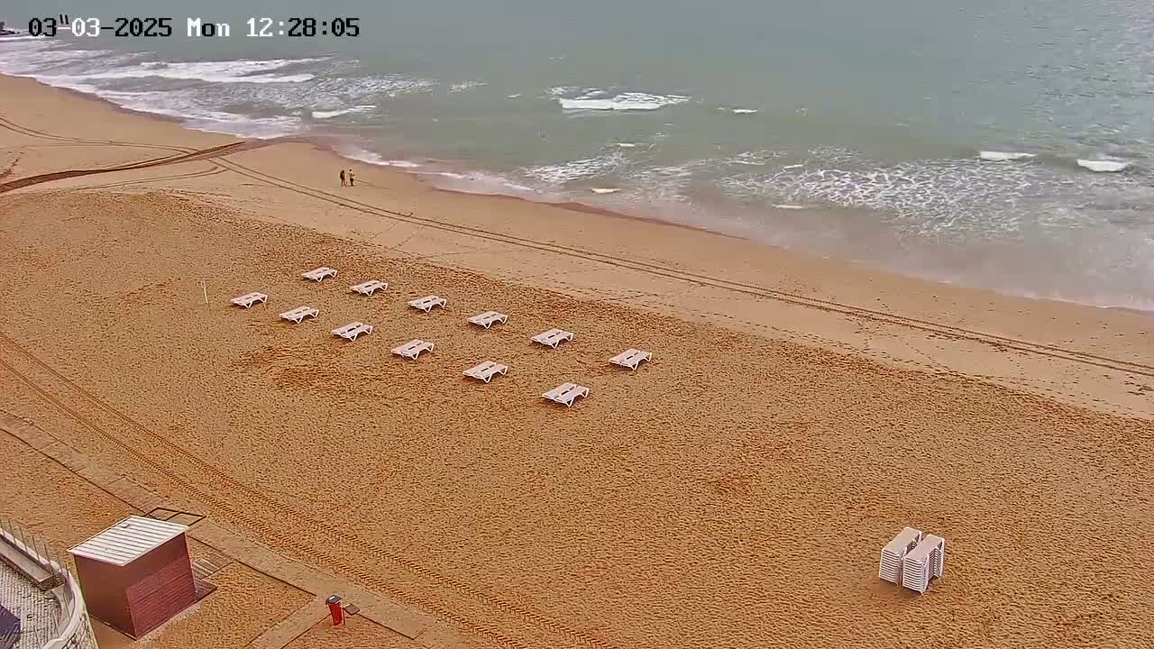 An overhead view shows a sandy beach with several empty lounge chairs arranged in rows, a small structure near the edge, and a calm ocean with small waves in the background.