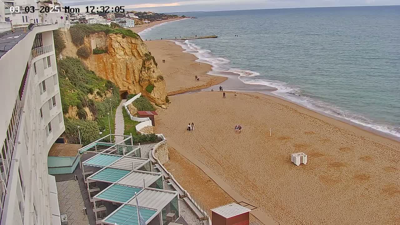 A sandy beach with a few people on it stretches along a calm ocean under an overcast sky, adjacent to a tall, curved white building and a steep, reddish-brown cliff.