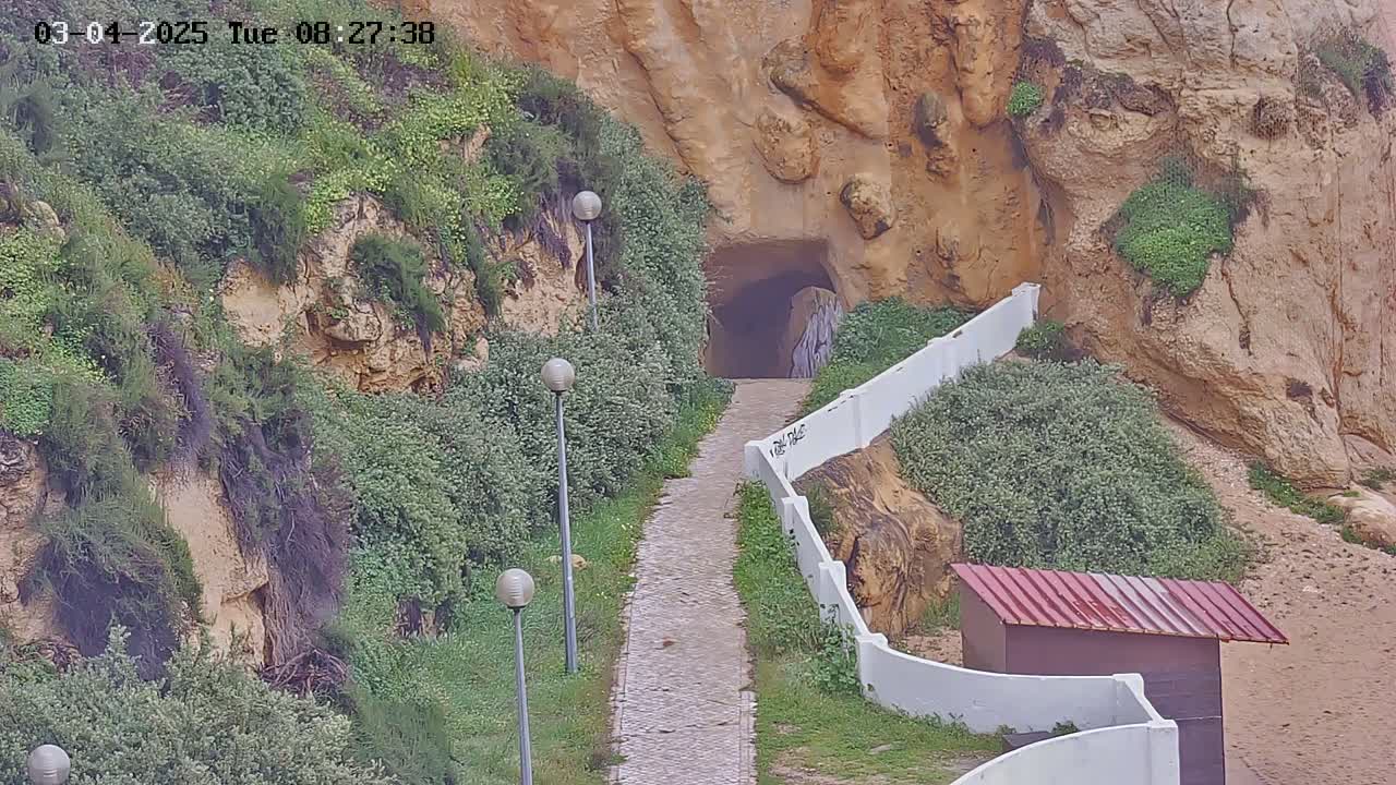 A paved pathway leads to a tunnel carved into a tan cliffside, flanked by low white walls and vegetation, under a cloudy sky.