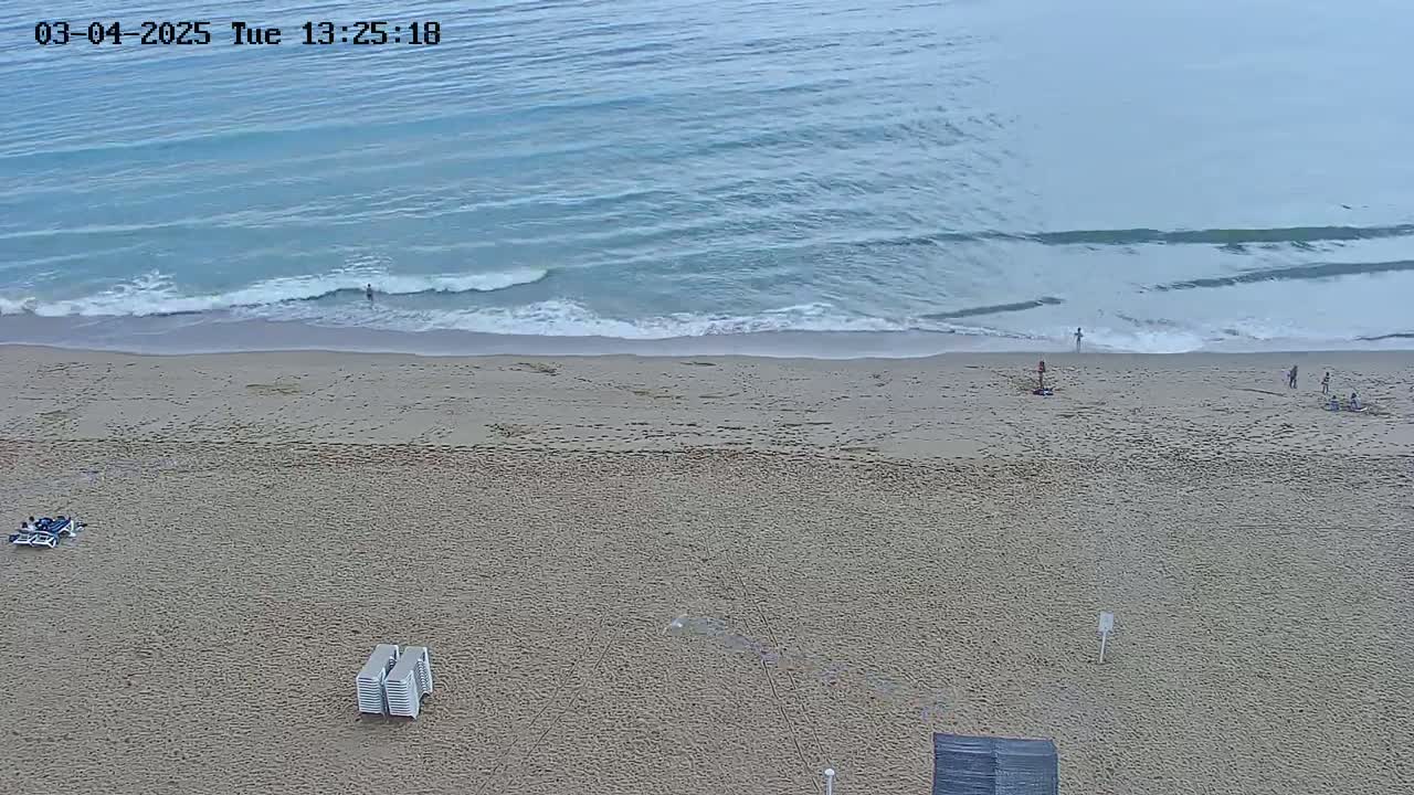 A mostly empty sandy beach with a few people and some beach chairs is shown, bordered by a calm, light-blue ocean with small waves.