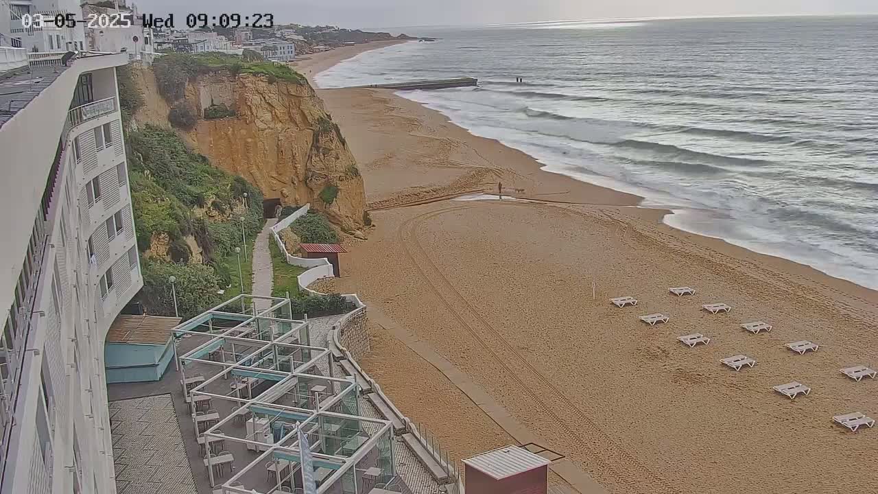 A sandy beach with several empty lounge chairs stretches below a high cliffside where a modern building stands, overlooking a moderately wavy ocean under an overcast sky.