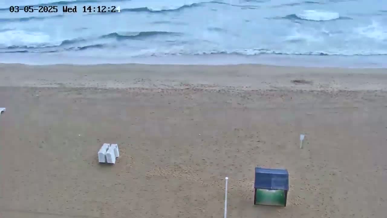 A sandy beach with a few pieces of beach furniture is shown with somewhat choppy waves in the ocean.