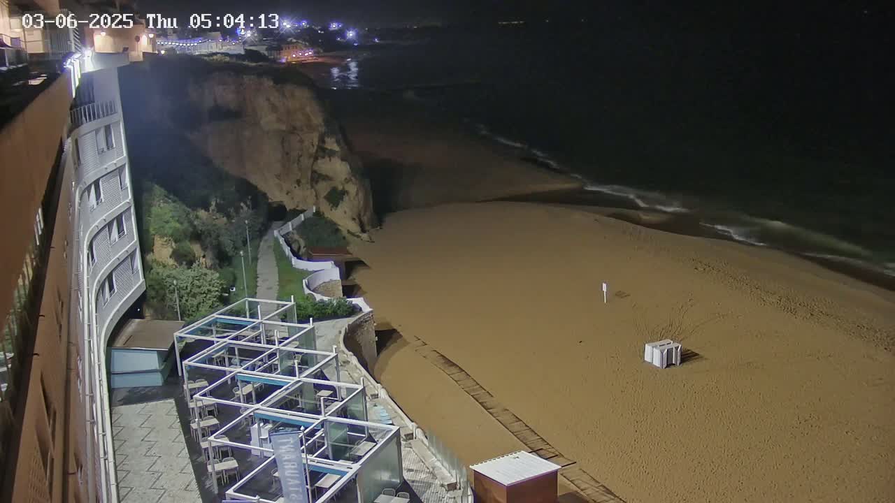 A sandy beach at night with gentle waves is adjacent to a cliffside building with outdoor seating.