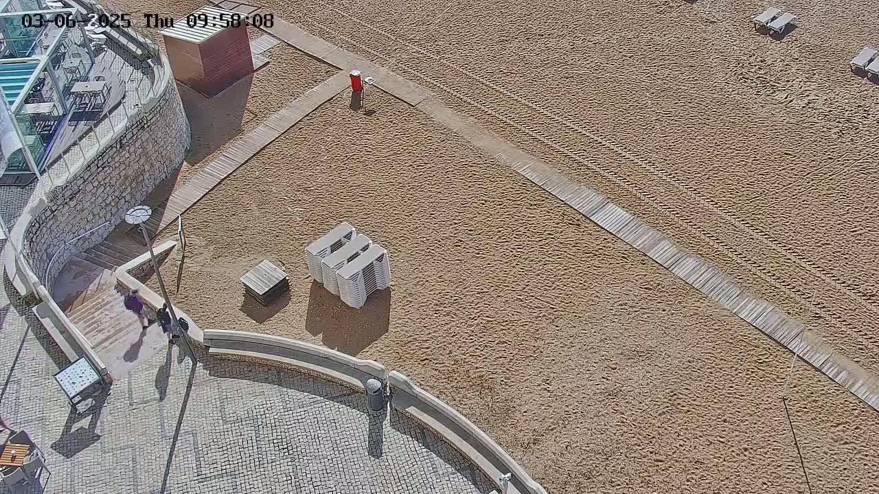 A sandy beach with wooden walkways, several stacked lounge chairs, and a few people walking near a stone staircase leading up to a building.