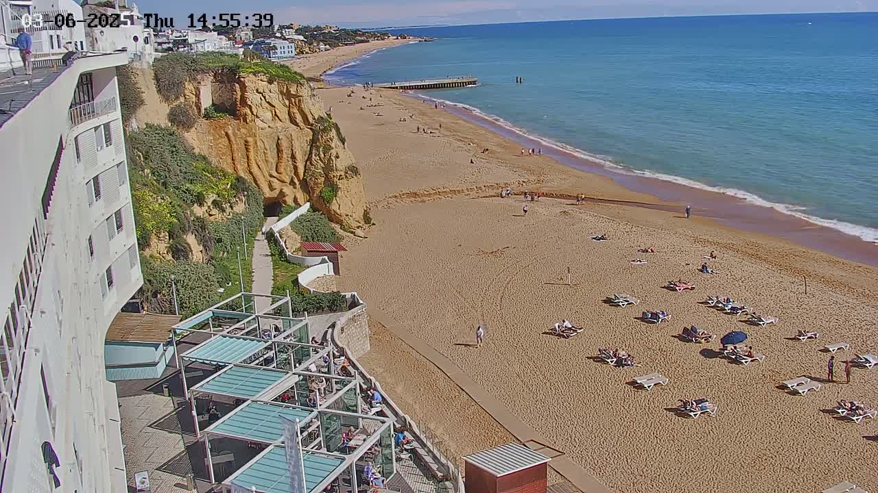 A sandy beach with sunbathers and lounge chairs stretches along a coastline under a clear sky, adjacent to a cliffside building with outdoor seating.