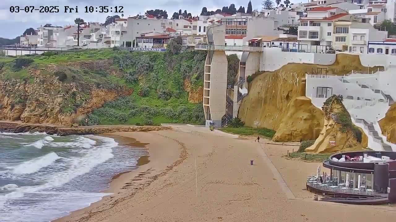 A sandy beach with moderate waves laps against a rocky shore where white buildings and a modern elevator structure are built into a cliff face under an overcast sky.