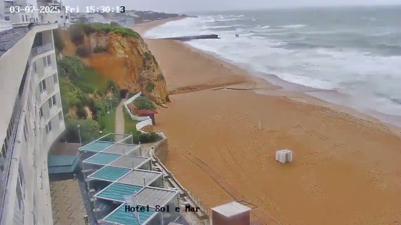 A sandy beach with choppy waves under an overcast sky is viewed from above, next to a tall white building and a cliff.