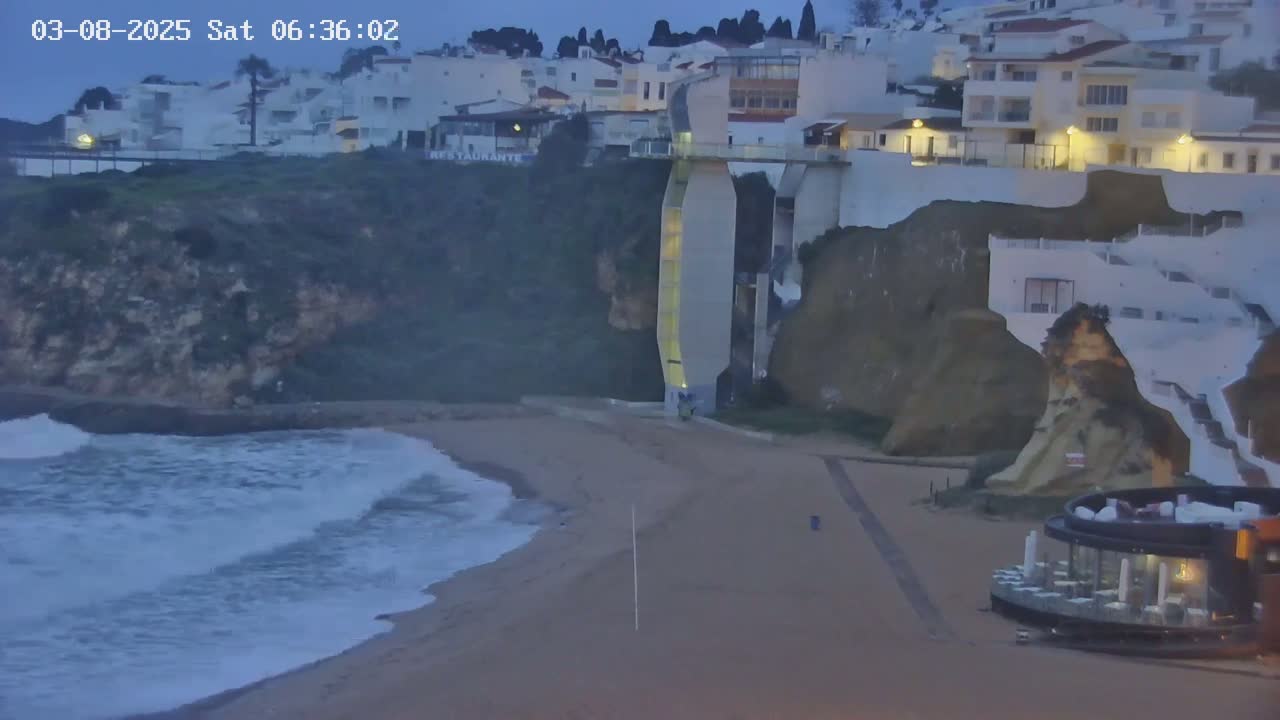 Waves crash on a sandy beach below a cliff with white buildings and a restaurant at dawn.