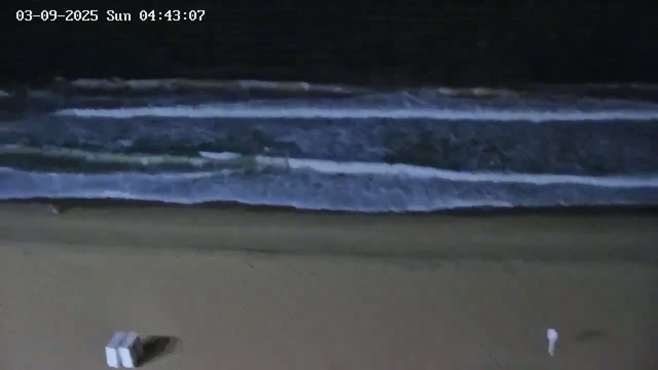 A dark beach at night shows several waves rolling onto the shore, with two objects resembling beach chairs near the bottom.