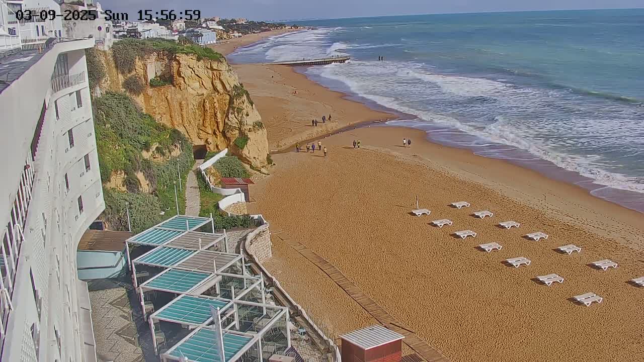 A sandy beach with a calm ocean, several people walking, and rows of empty lounge chairs, overlooks a white building on a cliffside.