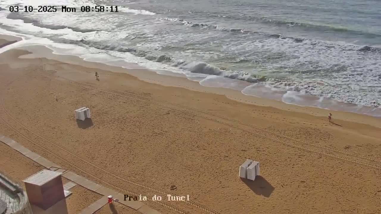An empty sandy beach with a few people and beach chairs, under a mostly sunny sky with moderate waves rolling onto the shore.
